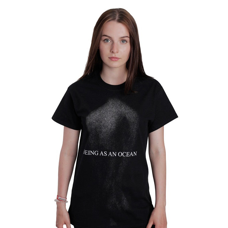 image of a white woman with brown hair wearing a black tee shirt on a white background. tee has a full body print in speckled grey of a persons nude back with the words being as an ocean written over the center in white.