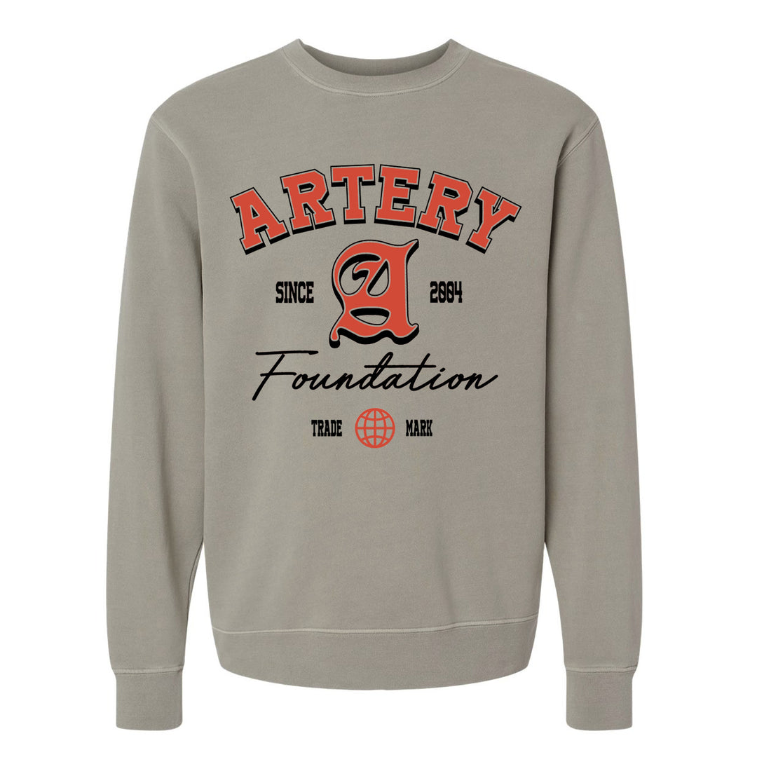 The Artery Foundation beige midweight pigment-dyed crewneck features bold red and black "ARTERY Foundation Since 2004" text, a large stylized A, and a small globe graphic above "TRADE MARK.