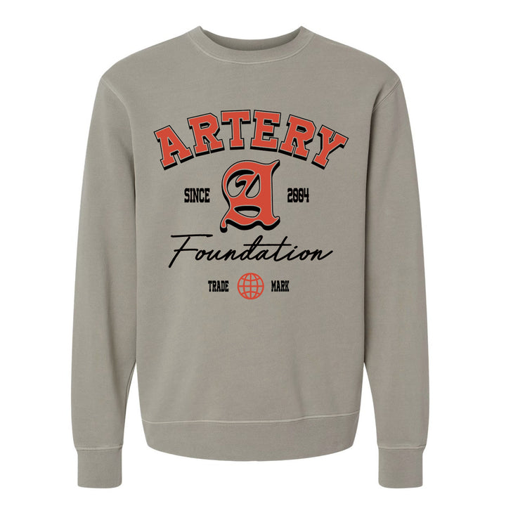 The Artery Foundation beige midweight pigment-dyed crewneck features bold red and black "ARTERY Foundation Since 2004" text, a large stylized A, and a small globe graphic above "TRADE MARK.
