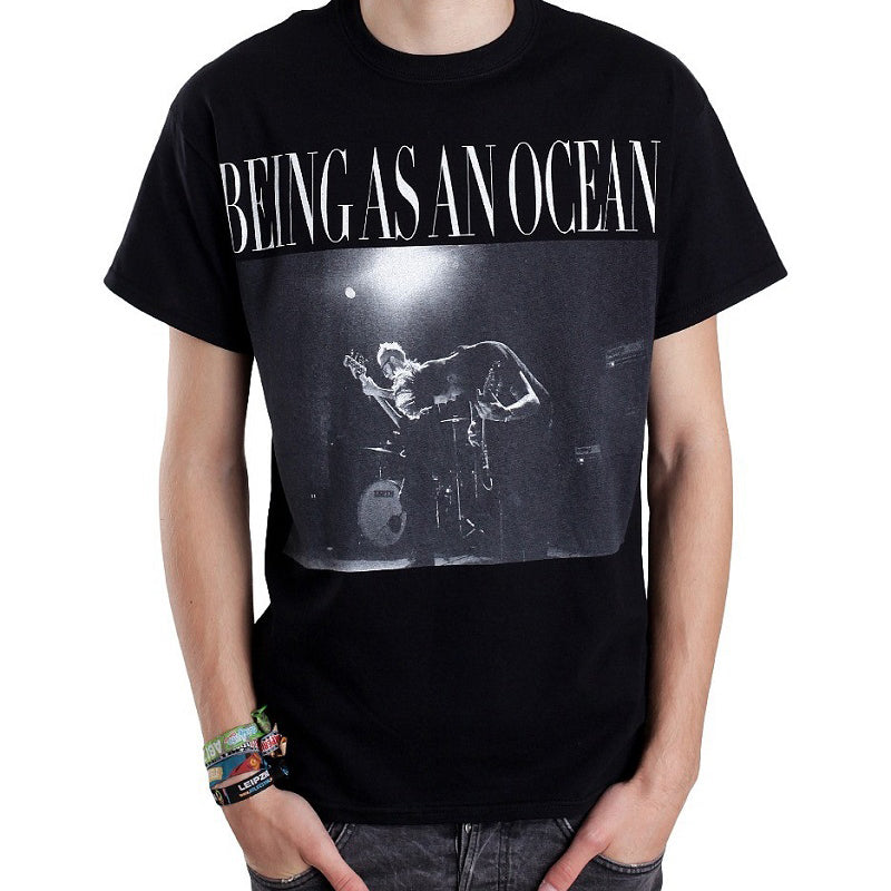 image of a white man with his hands in pants pockets wearing a black tee shirt on a white background. tee has full chest print in white that says being as an ocean at the top, with a photograph of two guys in the band playing guitar