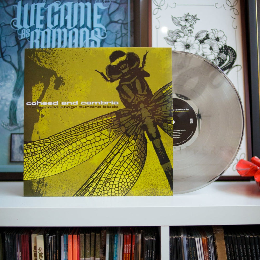 A transparent black Coheed and Cambria vinyl, The Second Stage Turbine Blade, with a dragonfly cover rests on a white shelf in front of framed artwork.