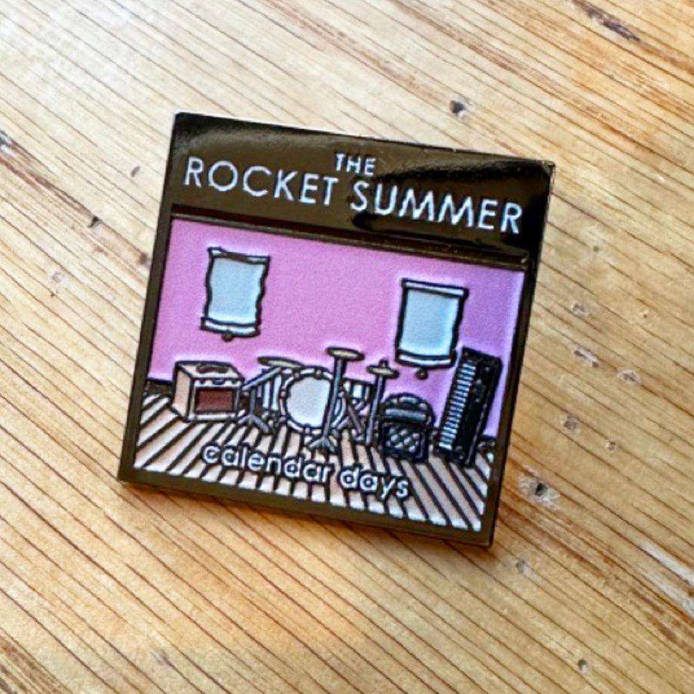 A 1 x 1 square enamel pin by The Rocket Summer, showing a cartoon drum set and music gear in a pink room with two windows. The top reads THE ROCKET SUMMER with calendar days below, displayed on wood.