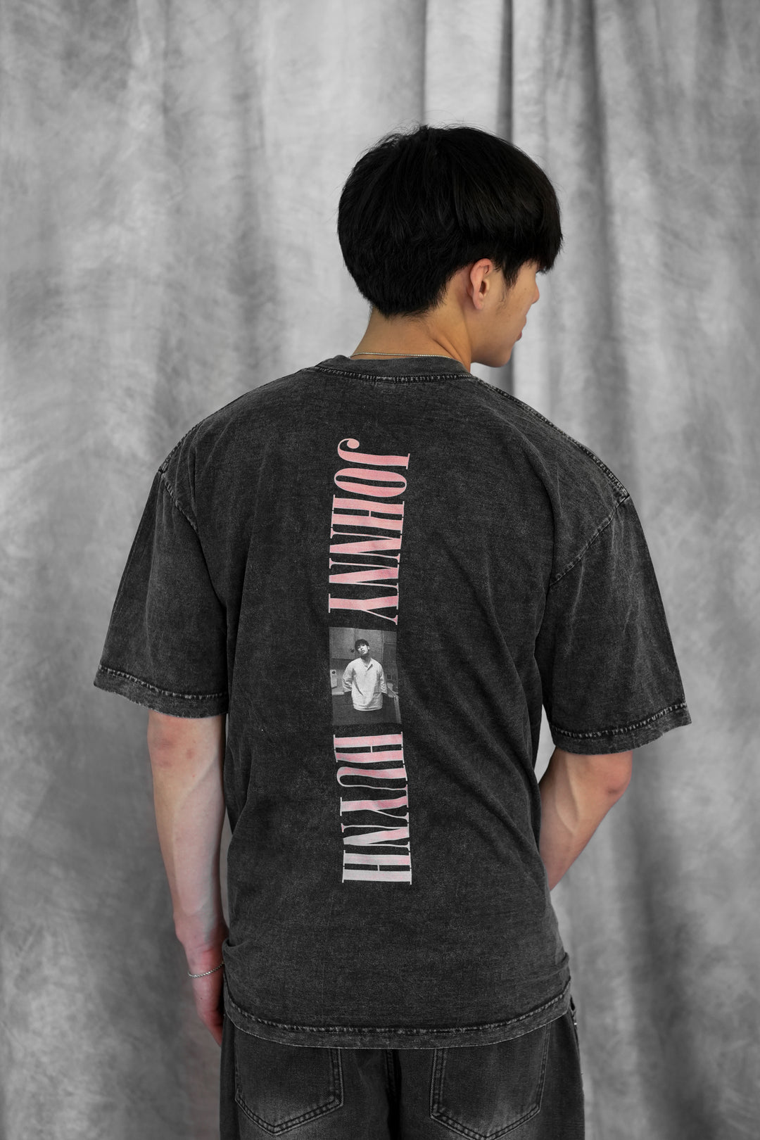 A person with short dark hair wears a faded black unisex mineral wash Johnny Huynh t-shirt, featuring the brand name and a small photo printed vertically on the back, standing against a gray, textured backdrop.