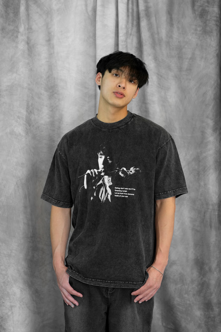 A young man with black hair stands before a gray textured backdrop, wearing a Johnny Huynh faded black mineral wash t-shirt with a black-and-white image of someone holding a microphone, paired with dark jeans.