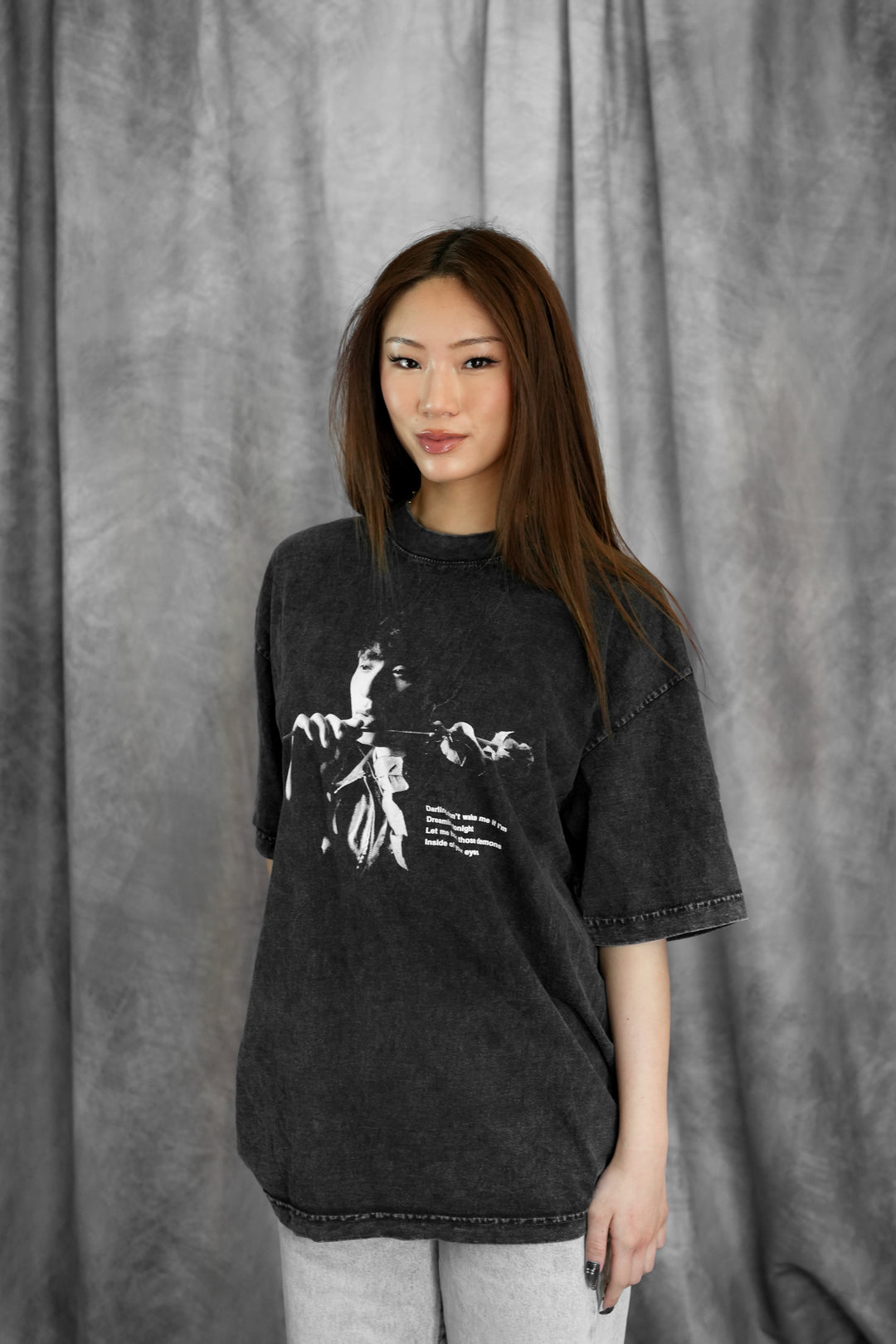 A young woman with long brown hair stands before a gray textured backdrop, wearing a Johnny Huynh unisex relaxed fit mineral wash graphic t-shirt and light gray pants. She looks at the camera with a slight smile.