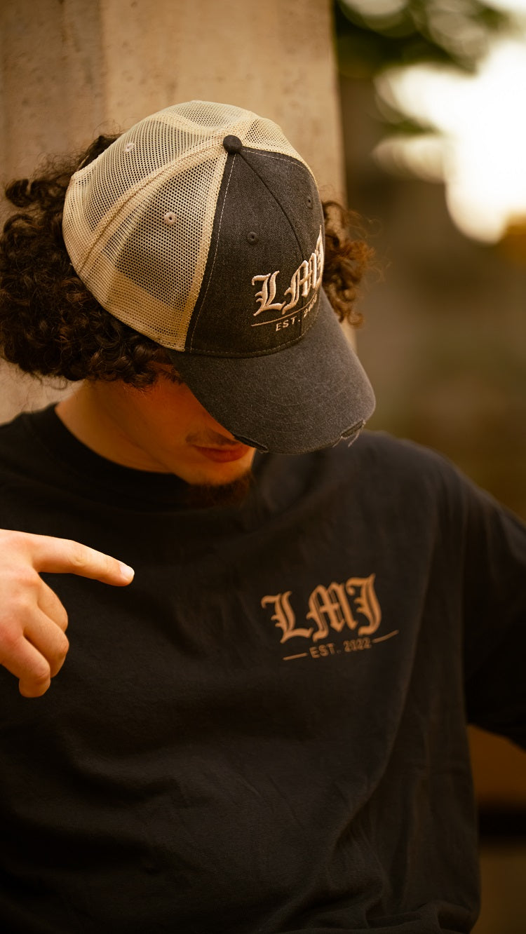 A person wearing a black and beige Lil Man J cap and a black t-shirt, both with "LMJ EST. 2022" text, points at the premium ring-spun cotton tee while looking down. The background is blurred.