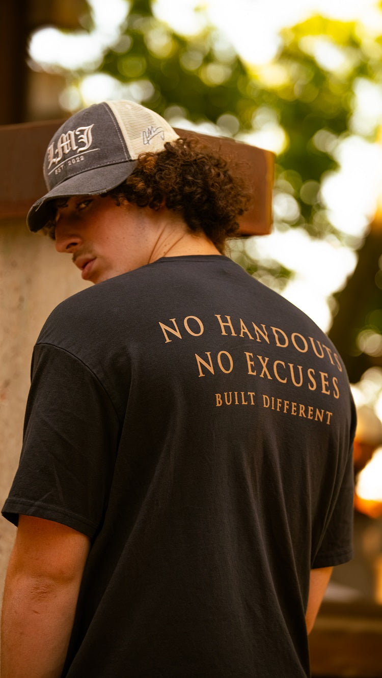A person in a cap and a black Lil Man J ring-spun cotton T-shirt that says "NO HANDOUTS NO EXCUSES BUILT DIFFERENT" stands outdoors, glancing over their shoulder with sunlight and trees behind them.