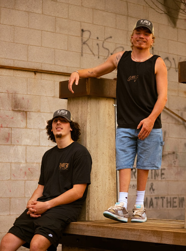 Two young men pose outside by a graffiti wall. One sits on a ledge in a black tee, shorts, and a Lil Man J cap. The other stands, leaning on a pillar in a black tank top, denim shorts, and an embroidered Lil Man J hat.
