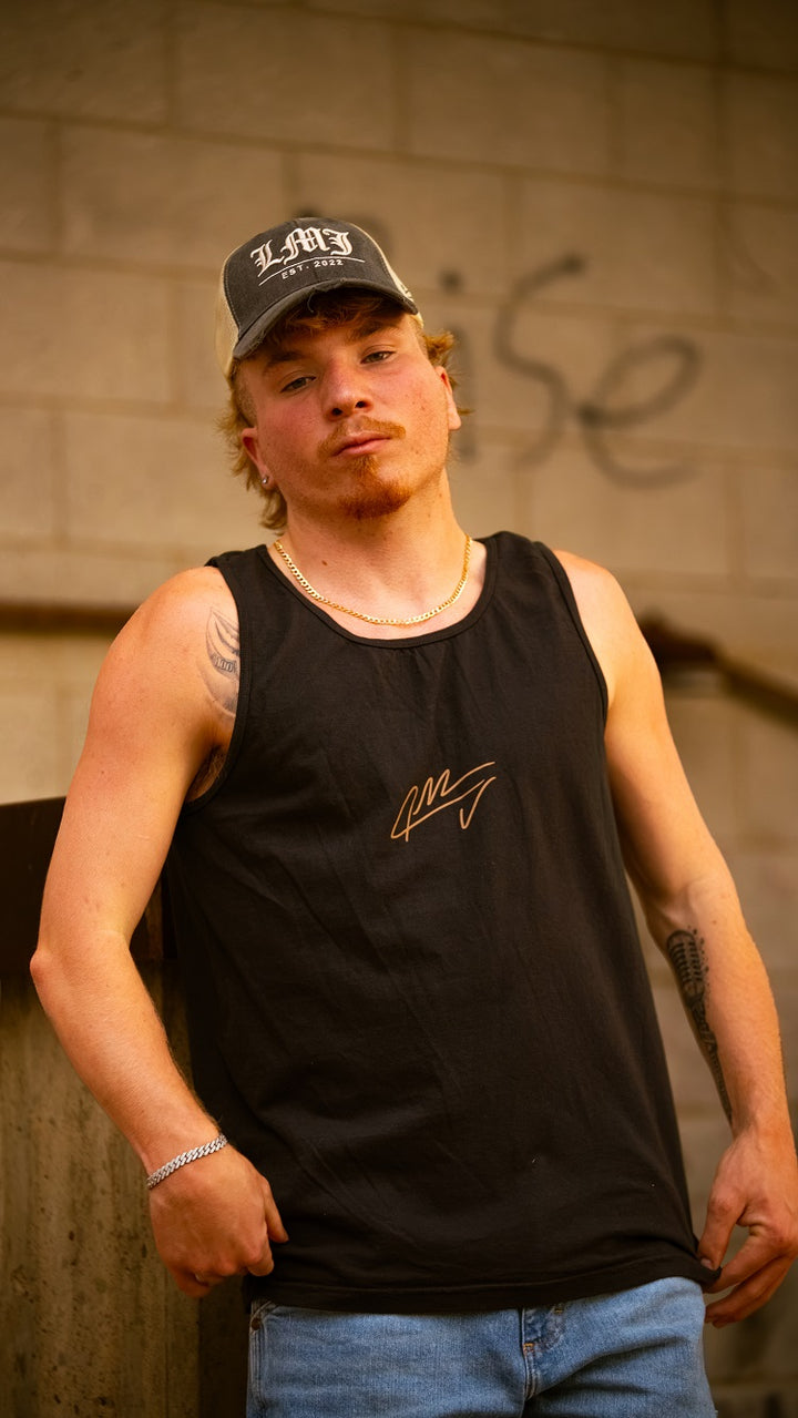 A young man in a Lil Man J black garment dyed tank top, blue jeans, and a gray cap leans against a concrete structure, showing off his arm tattoos and chain necklace. A graffiti-covered brick wall forms the background.