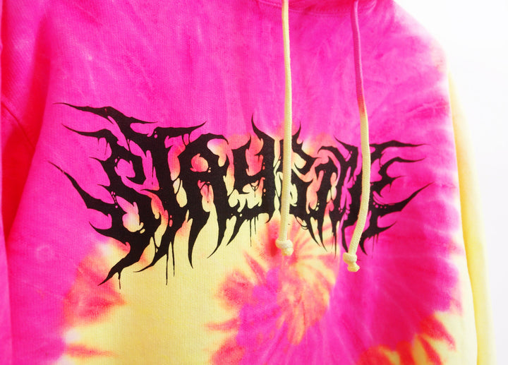 A pink and yellow Fluorescent Swirl tie-dye hoodie from Stay Sick Clothing features yellow drawstrings and bold black graffiti-style “STAY HOME” lettering across the chest—a standout streetwear piece.