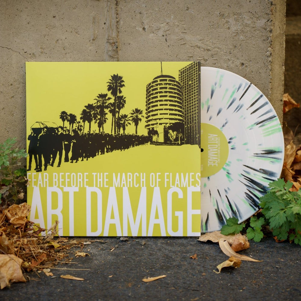 A limited edition Fear Before LP with a white, black, and mint green splatter is partially out of its yellow Art Damage cover, resting outdoors by a concrete wall with dry leaves and green plants nearby.
