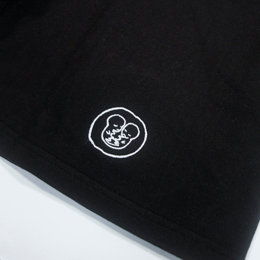Black fabric with a small white outlined graphic near the edge, depicting two cartoon-style faces within a circle. The design appears simple and minimalist, adding a playful touch to these AS Colour Stadium Shorts. Crafted from soft cotton sweat shorts material, they offer both comfort and style—perfect as understated embroidered shorts for everyday wear.