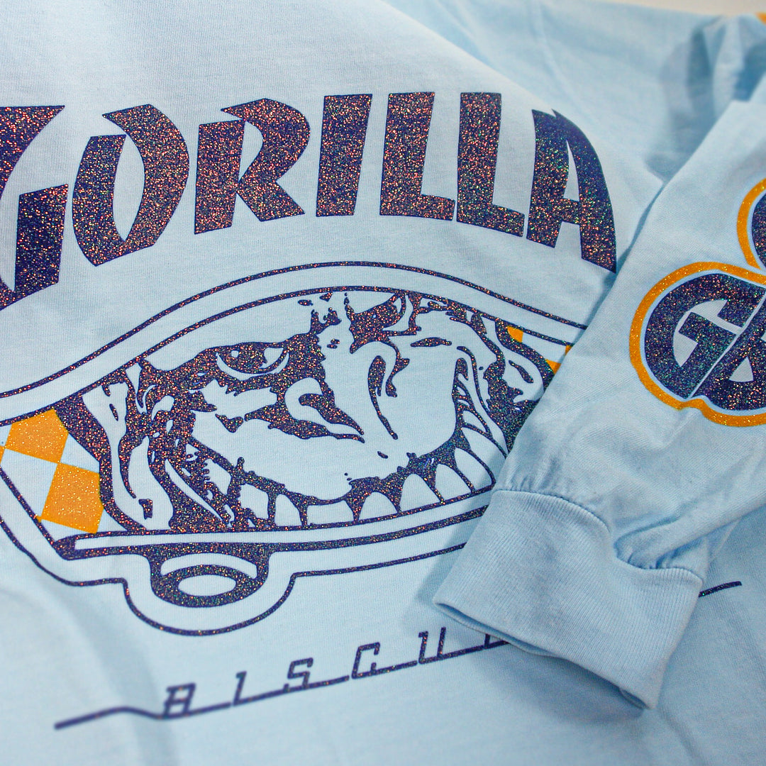 A light blue, relaxed fit Gorilla Biscuits shirt displays "GORILLA" in bold above a gorilla face and skateboard graphic, with a small GB patch on the long sleeve. Made from soft garment-dyed cotton with glittery accents.