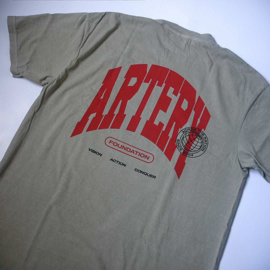 A beige, garment-dyed Artery Foundation Comfort Colors T-shirt with large red ARTERY text, a red oval with FOUNDATION, black VISION ACTION CONQUER text below, and a circular globe graphic on the right side.