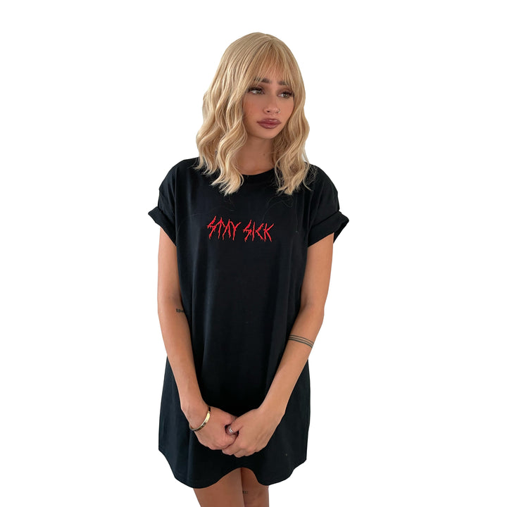 A woman with wavy blonde hair wears a loose black T-shirt dress by Stay Sick Clothing, featuring red metal-embroidered SAD SICK lettering, standing against a white background with her arms at her sides.
