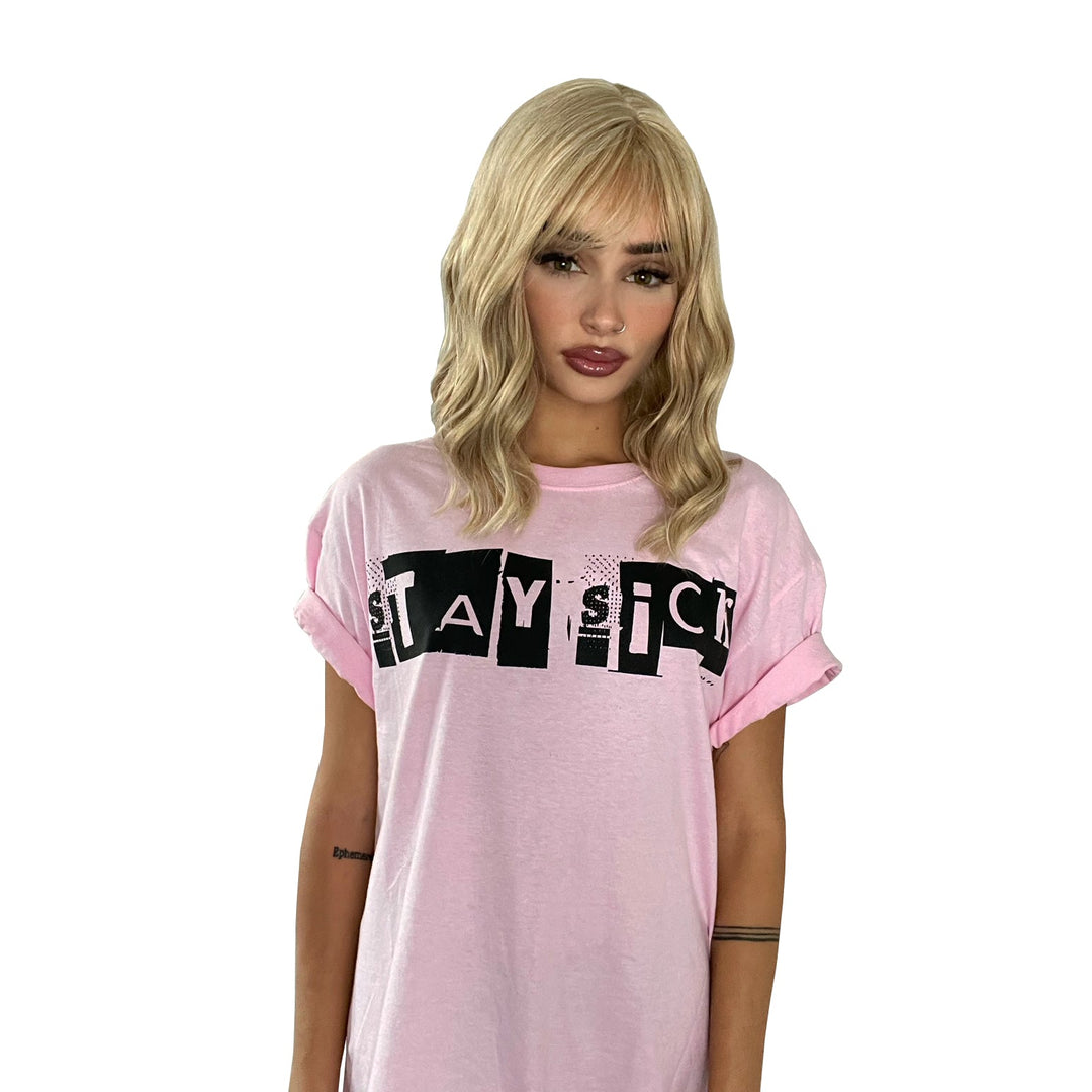 A woman with wavy blonde hair models a Stay Sick Clothing Ransom Pink T-Shirt featuring bold black STAY SICK lettering, posed against a plain white background.