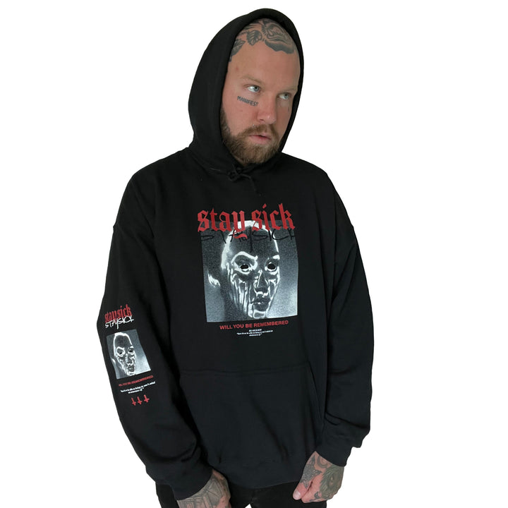 A man with face and hand tattoos wears a black Stay Sick Clothing hoodie featuring STAY SICK, a distressed face and Will You Be Remembered text, plus red and white sleeve graphics. He stands against a white background.