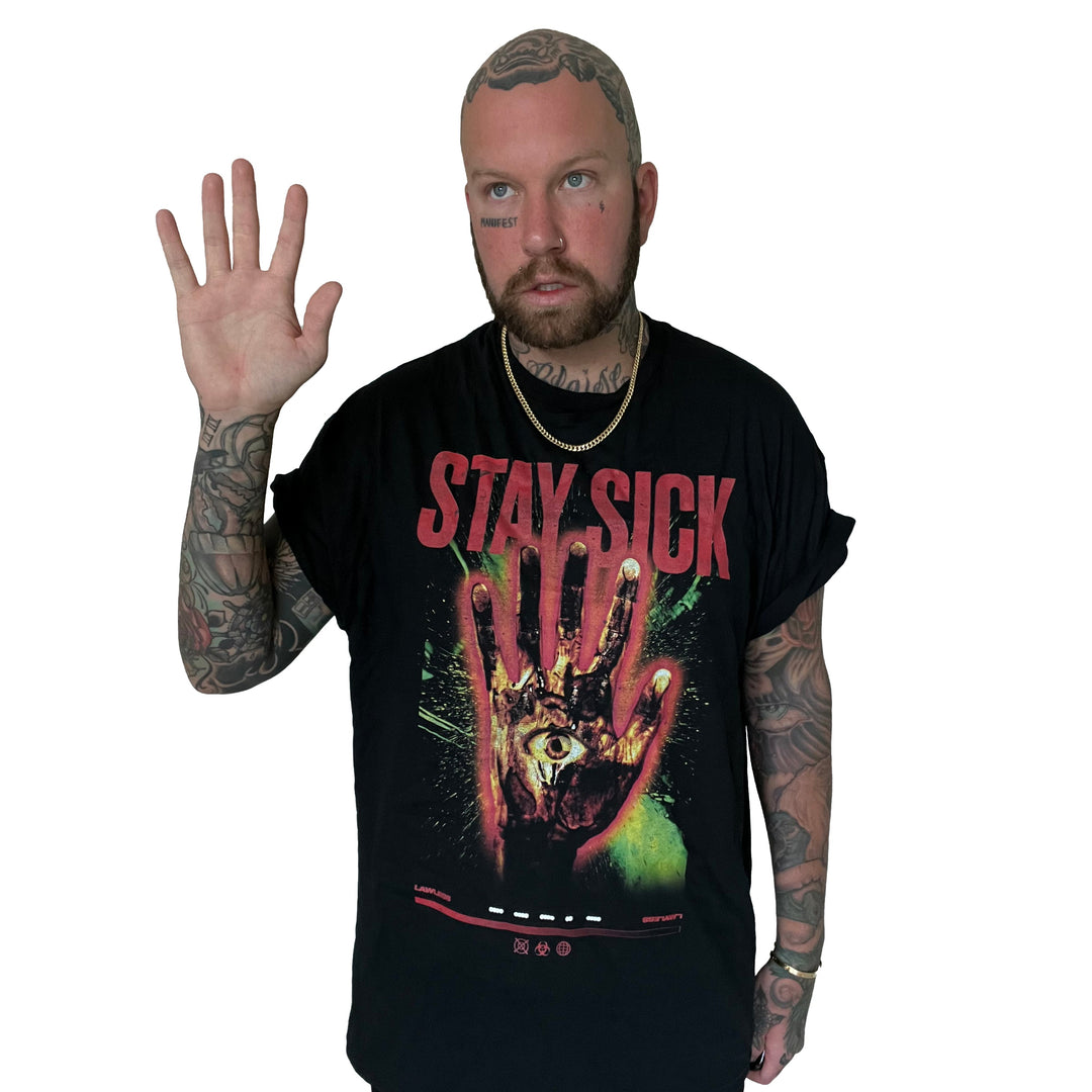 A bearded, tattooed man raises his right hand and looks aside, wearing a black Stay Sick Clothing T-shirt with a bold graphic of a hand featuring an eye in the center. The background is plain white.