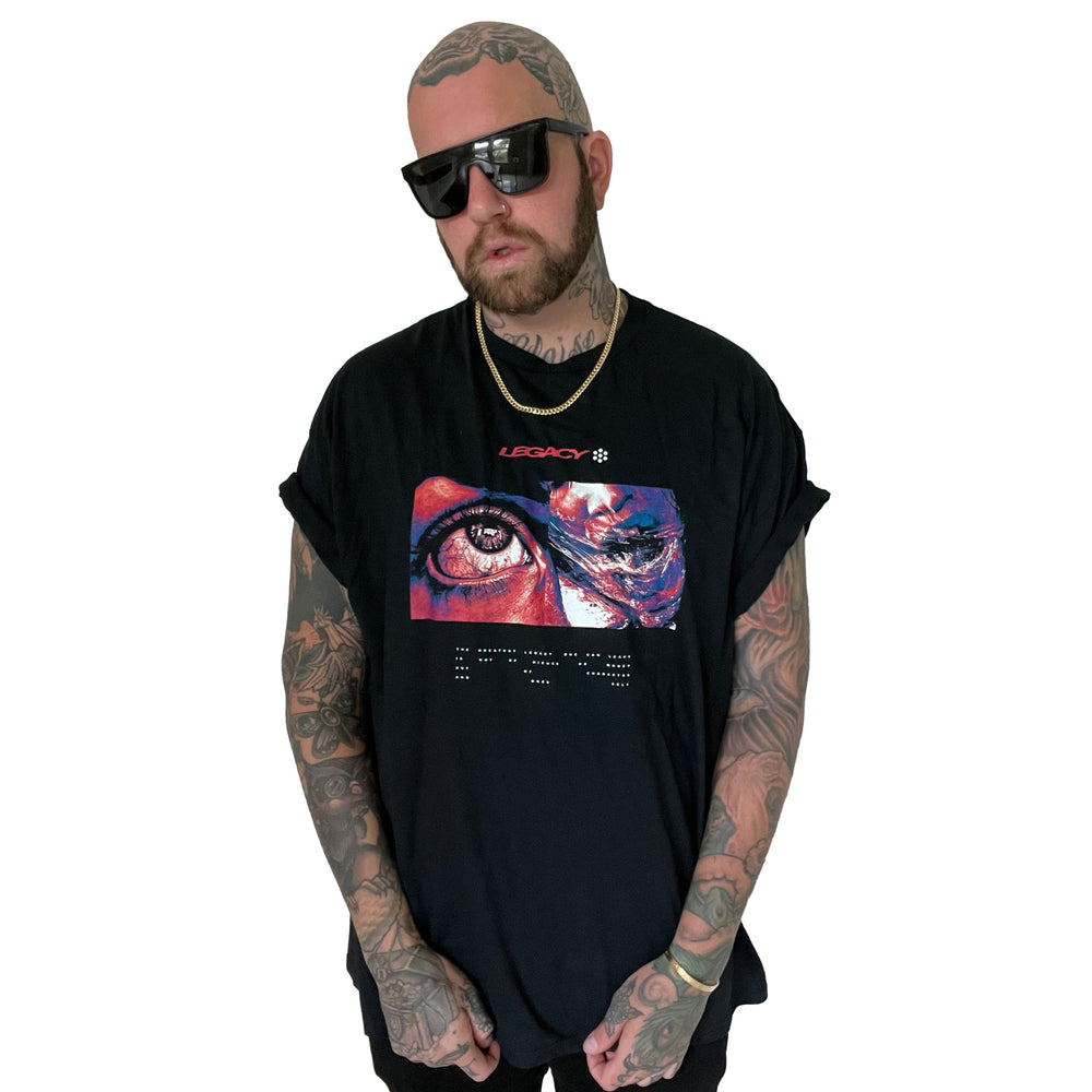 A bearded, tattooed man in sunglasses and a Stay Sick Clothing Legacy Black T-Shirt with a colorful eye and abstract art graphic, plus a gold chain, stands against a white background with hands in his pockets.