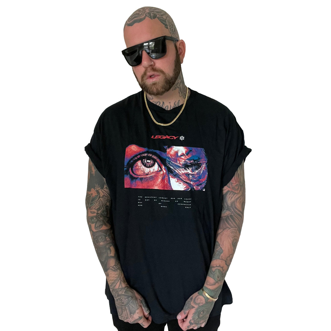 A bearded, tattooed man in sunglasses and a Stay Sick Clothing Legacy Black T-Shirt with a colorful eye and abstract art graphic, plus a gold chain, stands against a white background with hands in his pockets.
