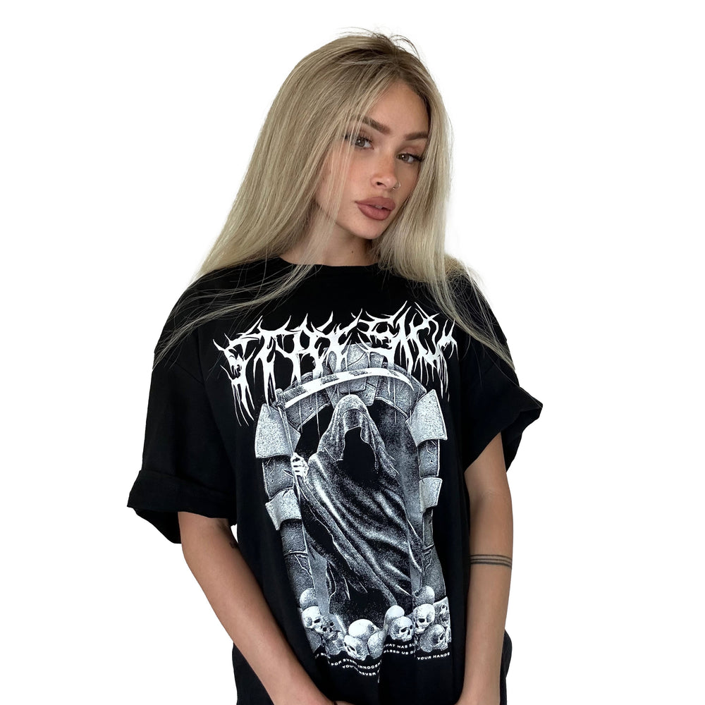 A woman with long blonde hair wears a Stay Sick Clothing Black T-Shirt with a death metal-style hooded reaper, skulls, and gothic text graphic, standing against a plain white background.