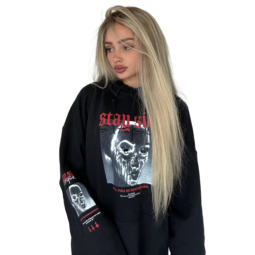 A young woman with long blonde hair models a Stay Sick Clothing black pullover with a graphic face and Will You Be Remembered in red text, standing against a plain white background and looking to the side.