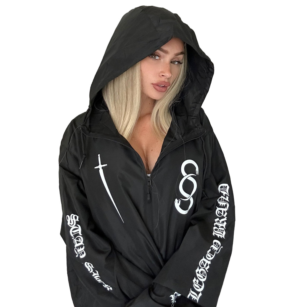 A woman with long blonde hair models a black zip-up jacket from Stay Sick Clothing, featuring white graphic designs and text on the sleeves and chest, against a plain white background.