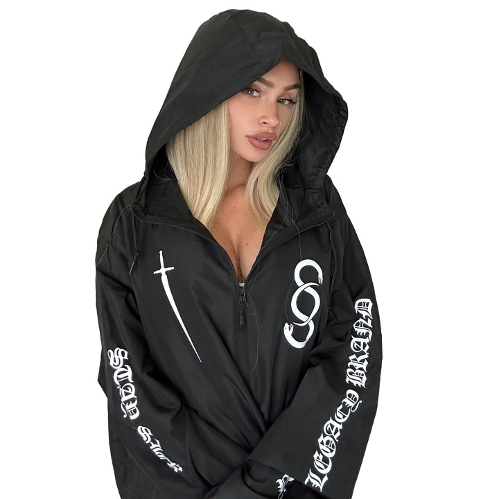 A woman with long blonde hair models a black zip-up jacket from Stay Sick Clothing, featuring white graphic designs and text on the sleeves and chest, against a plain white background.