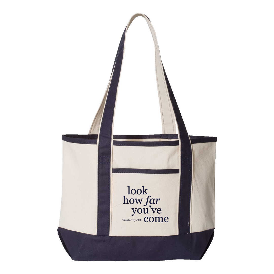 A cream-colored embroidered tote bag from Joshua Bassett with dark blue handles and trim. Features a front pocket printed in blue with the words "look how far you've come.