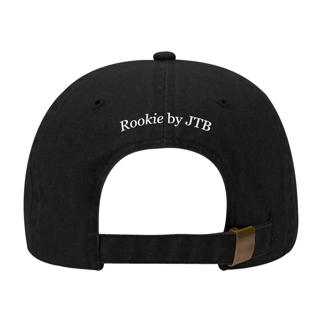 Black baseball cap viewed from the back, with white embroidered "Rookie by JTB" text above an adjustable strap and brass buckle. A Joshua Bassett piece inspired by OTTO Caps and classic dad hats for a timeless vibe.