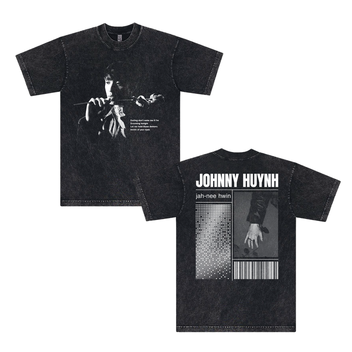 Black unisex garment-dyed cotton T-shirt by Johnny Huynh. Front: grayscale image of a person holding a flower, white text. Back: large JOHNNY HUYNH name with pronunciation, hand with flower, dotted pattern, and barcode design.