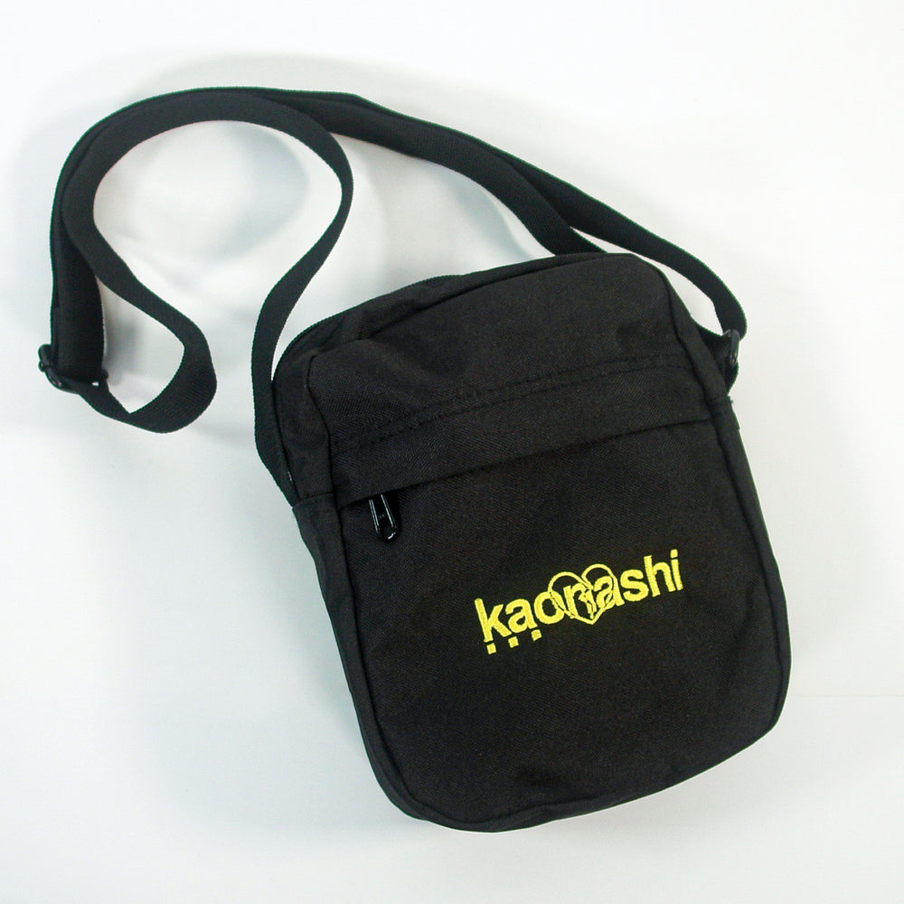 A compact black Kaonashi Flight Bag made from recycled polyester, with an adjustable strap, front zipper pocket, and striking yellow Kaonashi text and logo on the front.