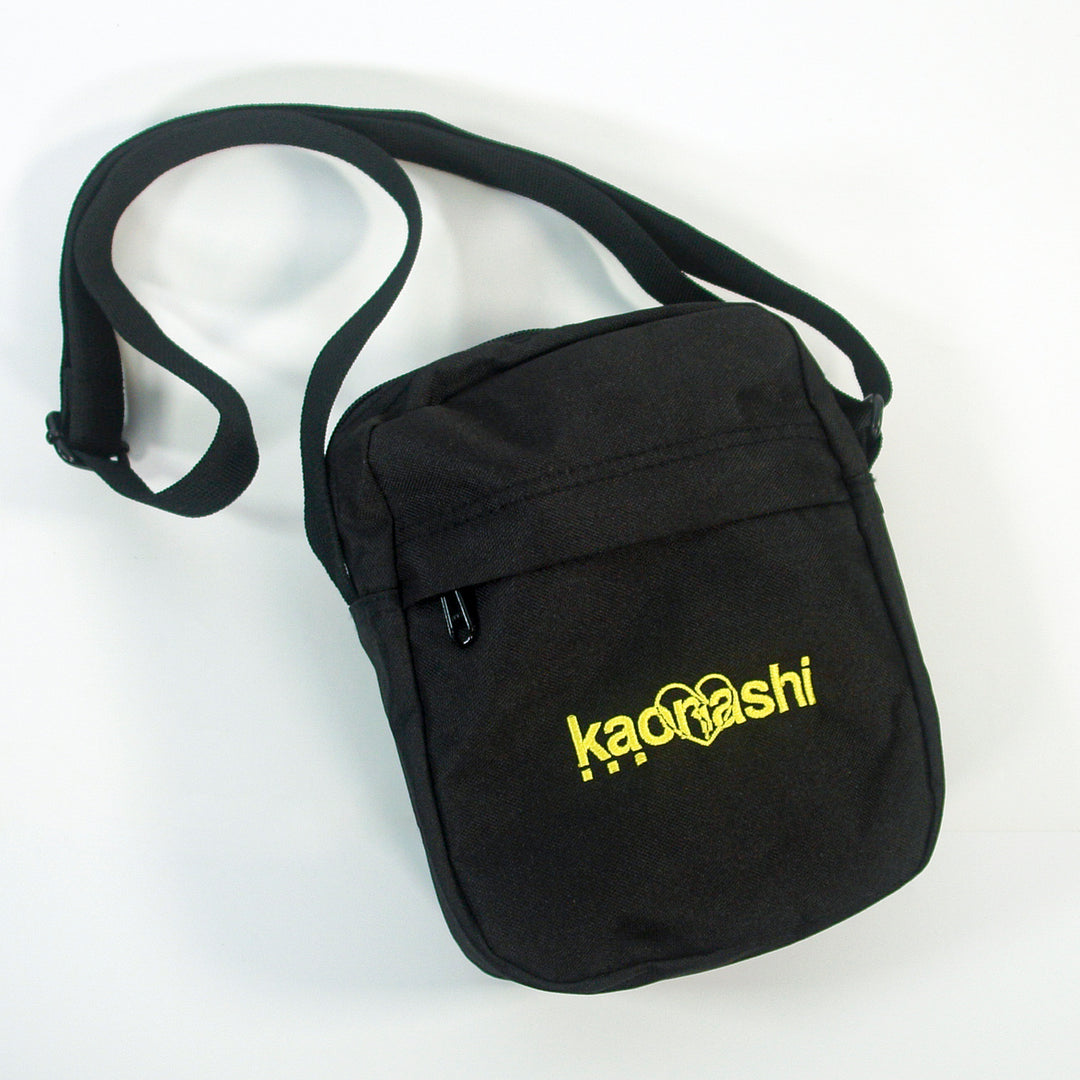 A compact black Kaonashi Flight Bag made from recycled polyester, with an adjustable strap, front zipper pocket, and striking yellow Kaonashi text and logo on the front.