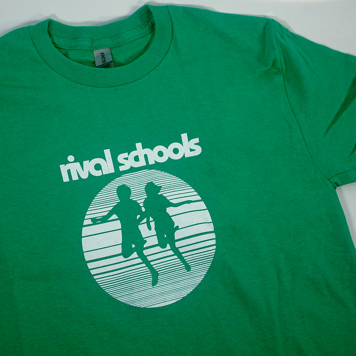 A green Rival Schools Gildan Heavy Cotton t-shirt with a classic fit, featuring a white graphic of two running figures in a striped circle and "rival schools" printed in bold lowercase letters above the design.