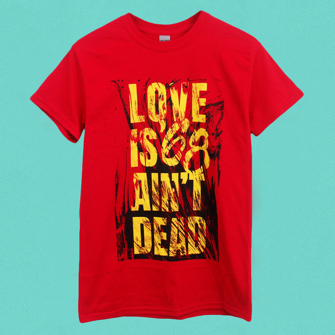 A classic-fit red '68 Gildan T-shirt in cotton polyester features bold yellow and black "LOVE ISNT DEAD" text on a blue textured background, with "ISNT" formed by red marks striking out "IS.