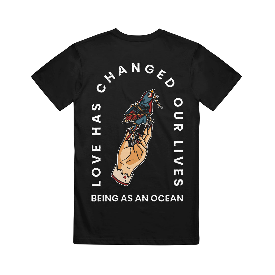 image of the back of a black tee shirt on a white background. the tee has a tan hand holding a blue and red bird tied in rope in the center with white text arched around that says love has changed our lives and being as an ocean written below.