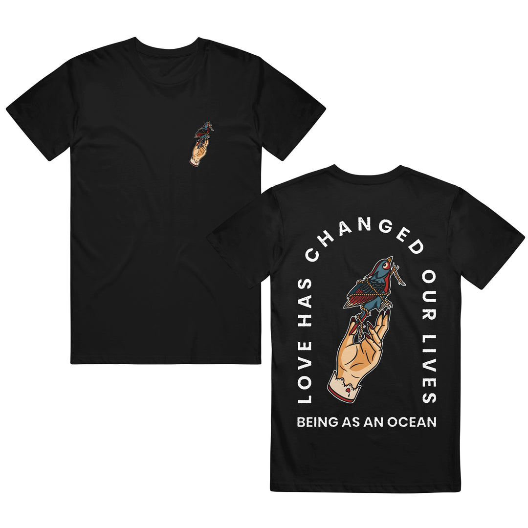 image of the front and back of a black tee shirt on a white background. the front of the tee is on the left and has a small print on the right chest of a tan hand holding a blue and red bird tied up in rope. the back of the tee is on the right and has a tan hand holding a blue and red bird tied in rope in the center with white text arched around that says love has changed our lives and being as an ocean written below.