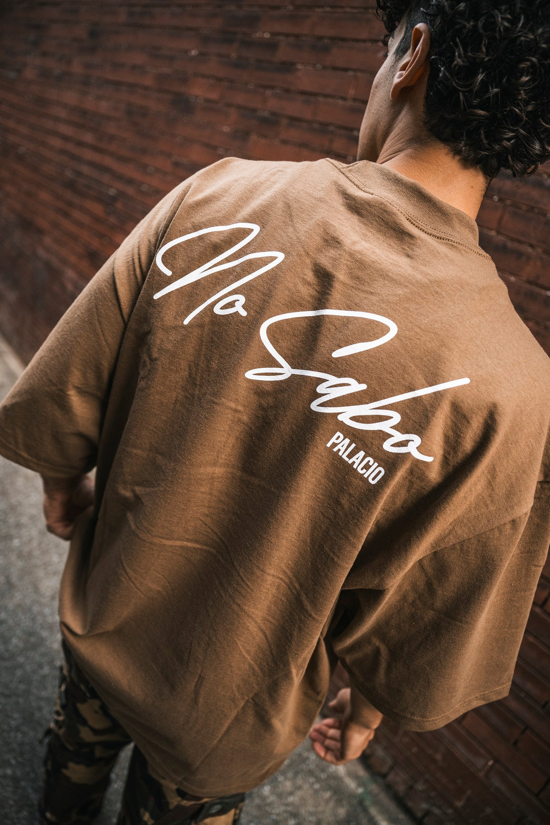 A person with curly hair faces away, wearing an oversized brown Micah Palace t-shirt made from 100% USA Cotton, featuring white "No Sabo PALACIO" script on the back, standing outdoors by a brick wall.