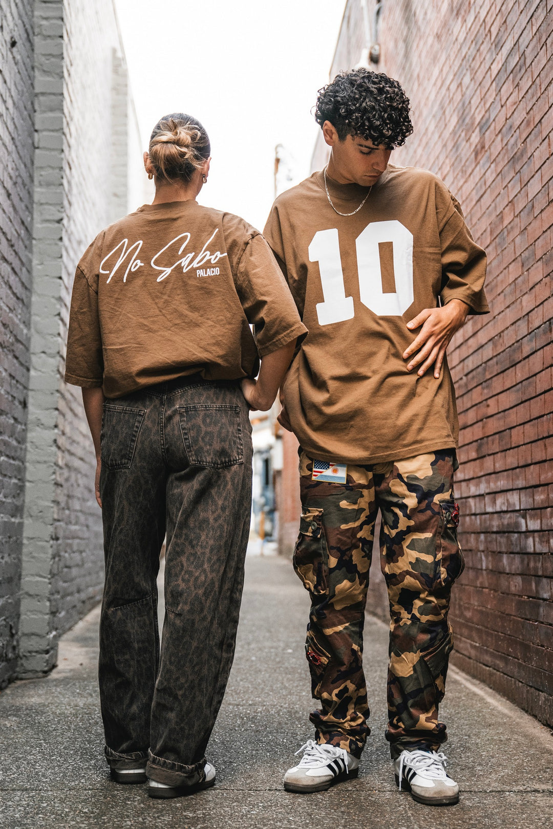 Two people stand in a narrow alley wearing brown Micah Palace Max Heavyweight T-Shirts with an oversized fit—one says No Sabo, the other 10. They keep it relaxed and casual by pairing the shirts with dark jeans and camo pants.
