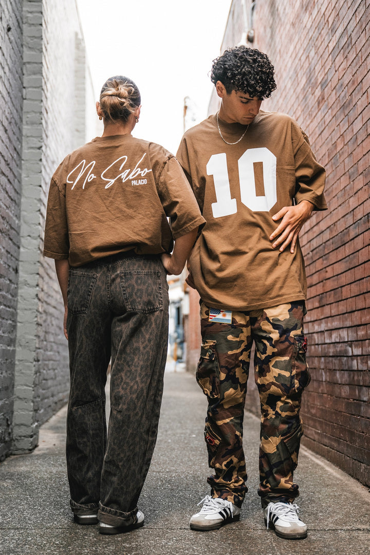 Two people stand in a narrow alley wearing brown Micah Palace Max Heavyweight T-Shirts with an oversized fit—one says No Sabo, the other 10. They keep it relaxed and casual by pairing the shirts with dark jeans and camo pants.