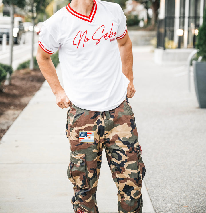 A person is outdoors, face unseen, wearing a white Micah Palace No Sabo shirt with red trim in moisture-wicking fabric and camouflage cargo pants with a USA flag patch.