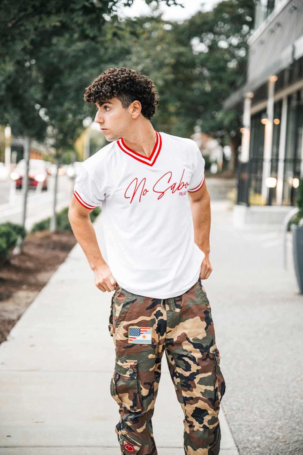 A young man stands on a sidewalk in a Micah Palace white No Sabo shirt with red trim, inspired by athletic soccer jerseys made from moisture-wicking Dryflex fabric, and camo pants with an American flag patch. He looks to his left in an urban setting.