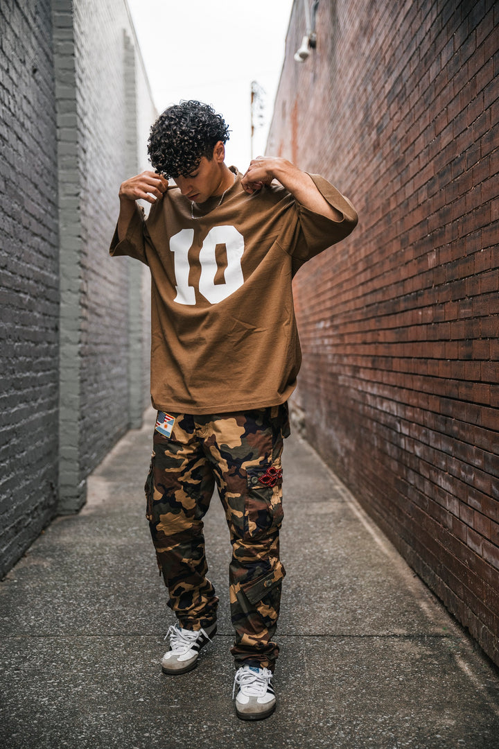 A person with curly hair stands in a narrow alley, adjusting a brown Micah Palace Max Heavyweight T-Shirt with the number 10. The oversized fit complements camo pants and gray sneakers for a stylish streetwear look.