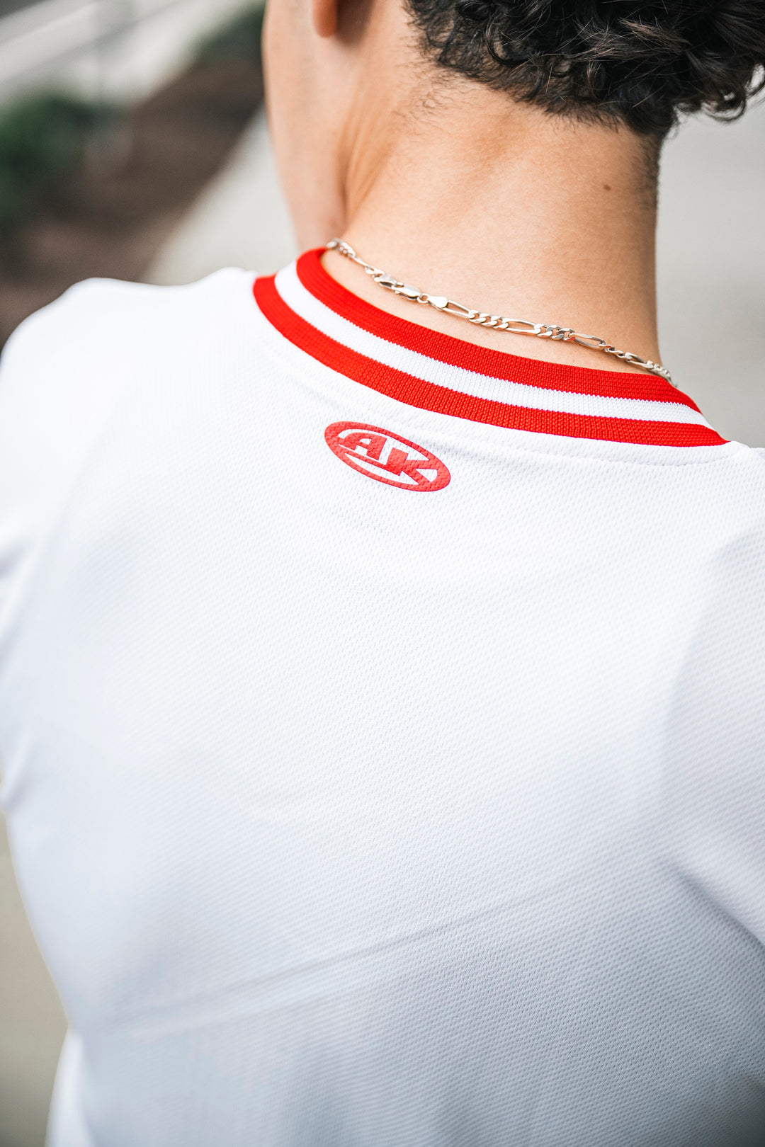 A person shown from behind wears a Micah Palace Soccer Jersey with a white body, red collar, small red logo on the back, and a gold chain necklace. The Moisture Wicking Dryflex fabric ensures optimal comfort.