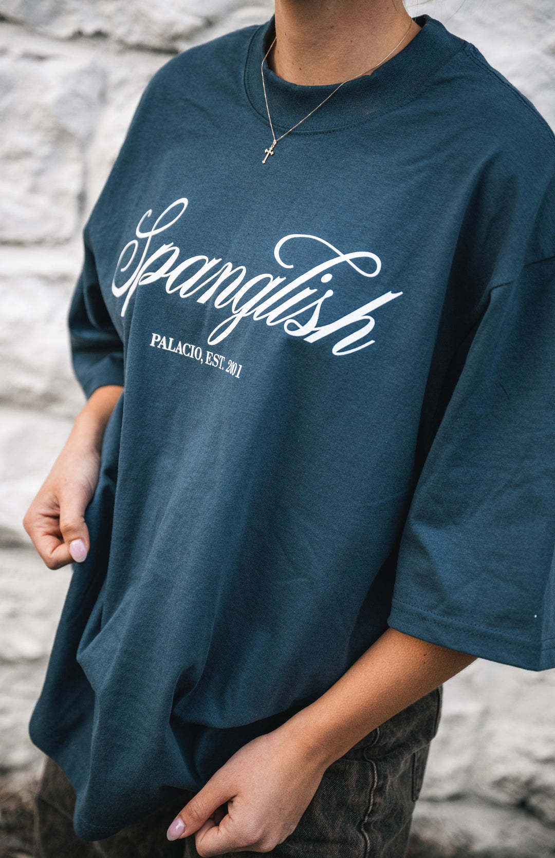 Someone wears an oversized dark blue t-shirt by Micah Palace, made from 100% USA cotton, with "Spanglish" and "PALACIO, EST. 201" in white script, paired with a cross necklace against a light stone wall.