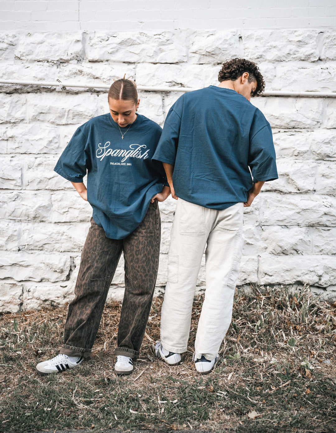 Two people stand side by side outdoors in front of a white stone wall, both wearing Micah Palace Max Heavyweight T-Shirts with an oversized fit. One faces forward with hands in pockets, the other faces away. Both wear casual pants and sneakers.
