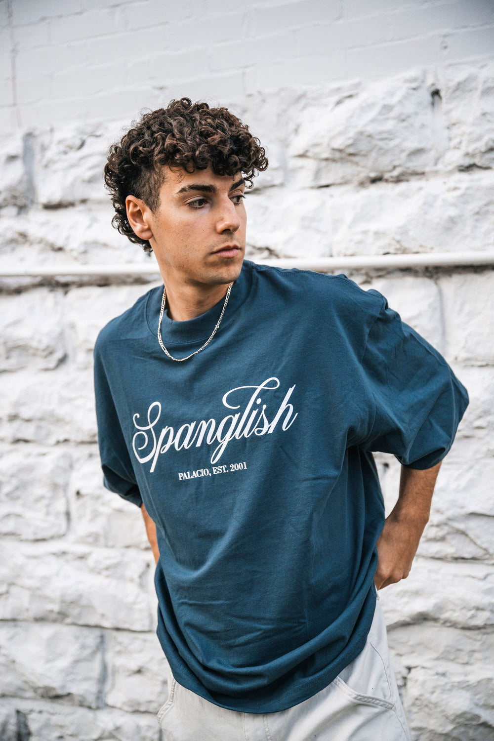 A young man with curly hair wears an oversized blue Micah Palace EST. 2001 T-shirt made from 100% USA Cotton and a chain necklace, standing by a white brick wall and gazing thoughtfully to the side.