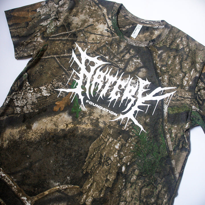 An Artery Foundation REALTREE camo t-shirt features "FOUNDATION" and an abstract white graphic across the chest. Made from soft ring-spun cotton, the shirt is shown laid flat on a white background.