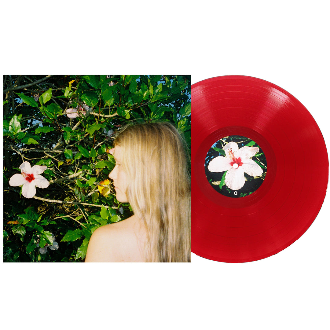 A red Oblivion vinyl record from Alice Phoebe Lou, featuring a hibiscus flower label, is displayed next to an album cover depicting Alice Phoebe Lou with her long blonde hair cascading as she stands before leafy green bushes adorned with white hibiscus flowers. Includes a download code for digital access.