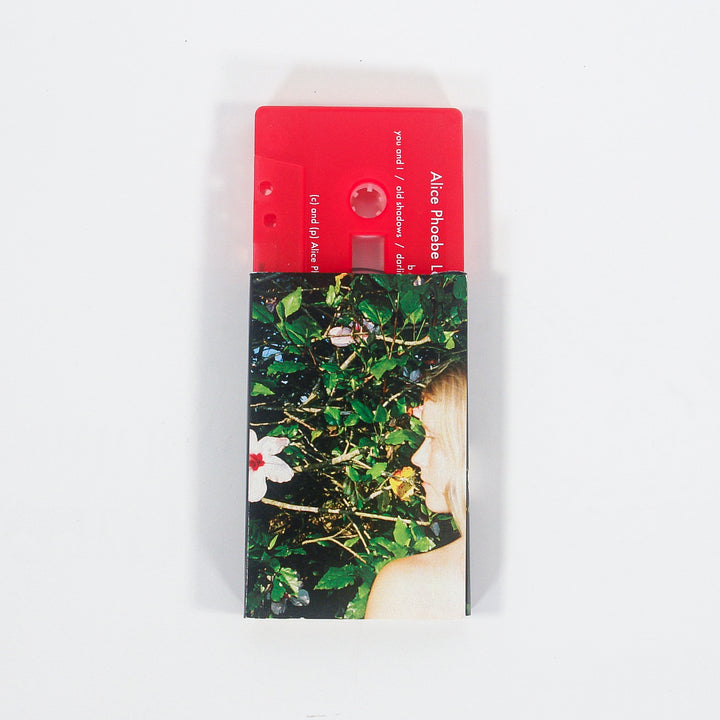A red cassette tape labeled Alice Phoebe partially slides out of a sleeve featuring a woman with blonde hair standing in front of green foliage and blooming flowers, capturing a nostalgic music cassette vibe that hints at the dreamy mood of Oblivion.